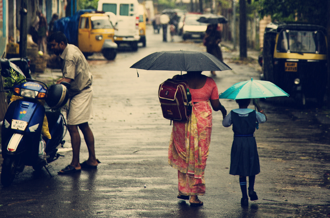 A mother in a sari walks her young daughter to school in the rain, both holding umbrellas
