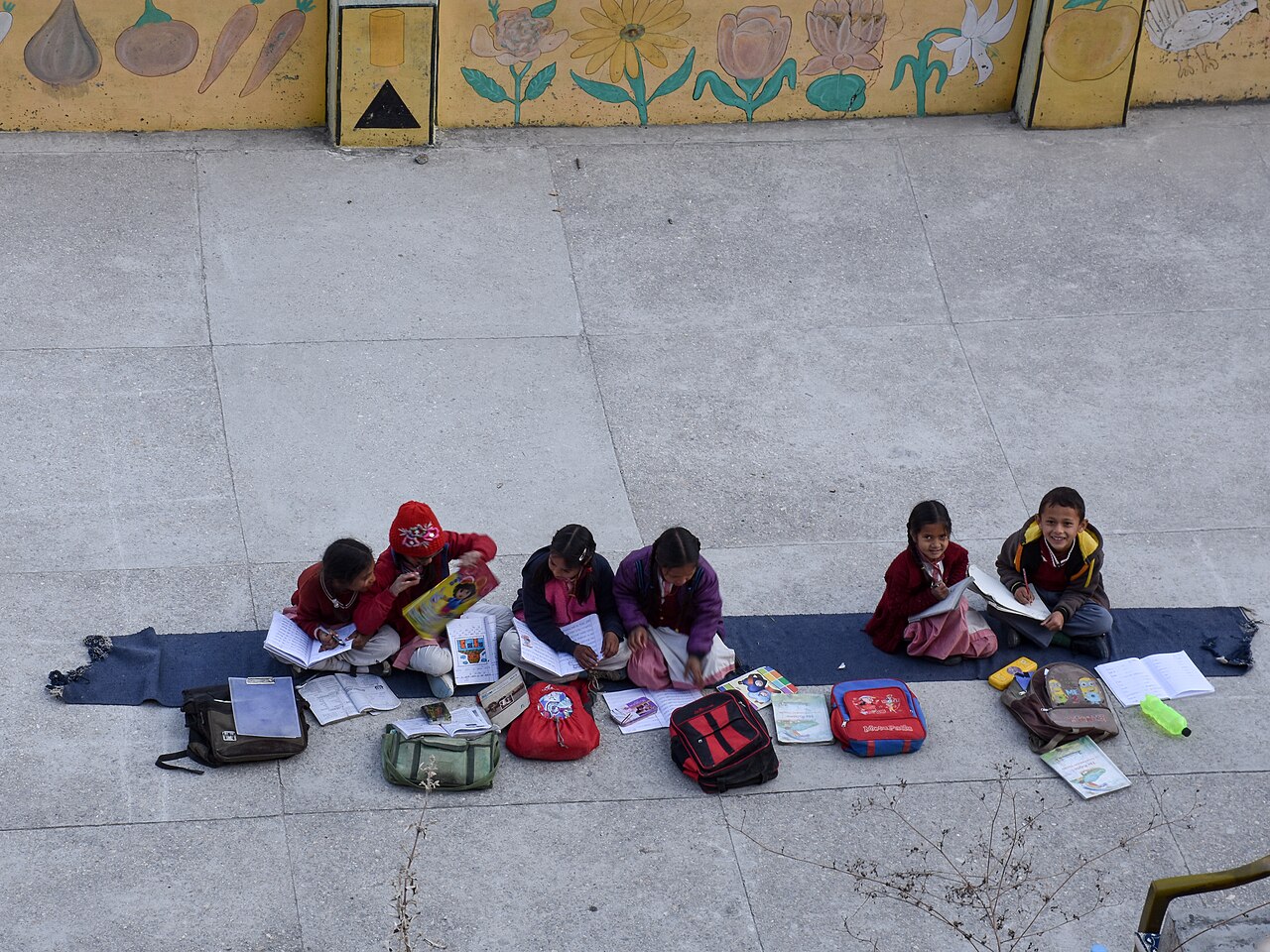 A group of school children sitting together on a mat, doing their homework with books and school bags around them